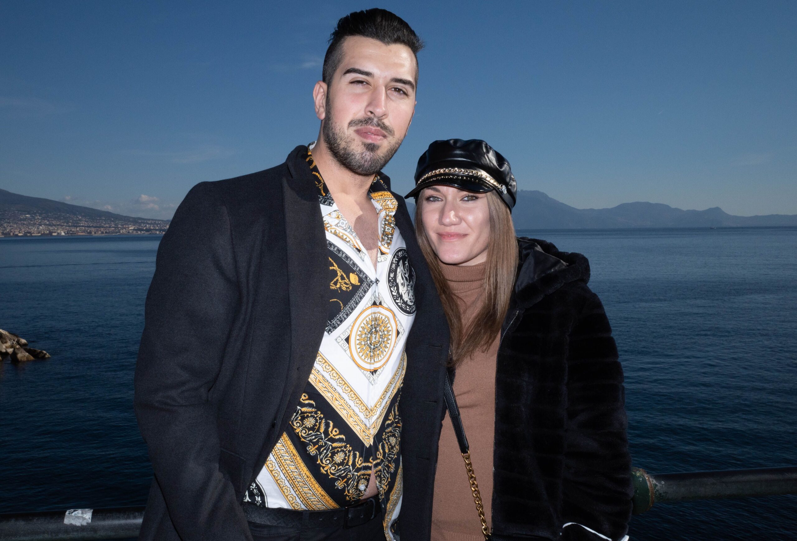 A happy couple stands by the ocean with a scenic mountain backdrop, enjoying their vacation.