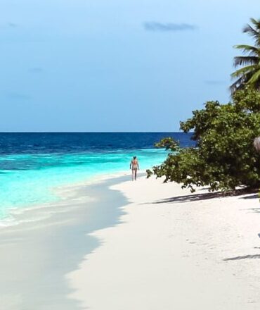 A woman strolling along a pristine tropical beach during the day, surrounded by clear blue waters and lush greenery.